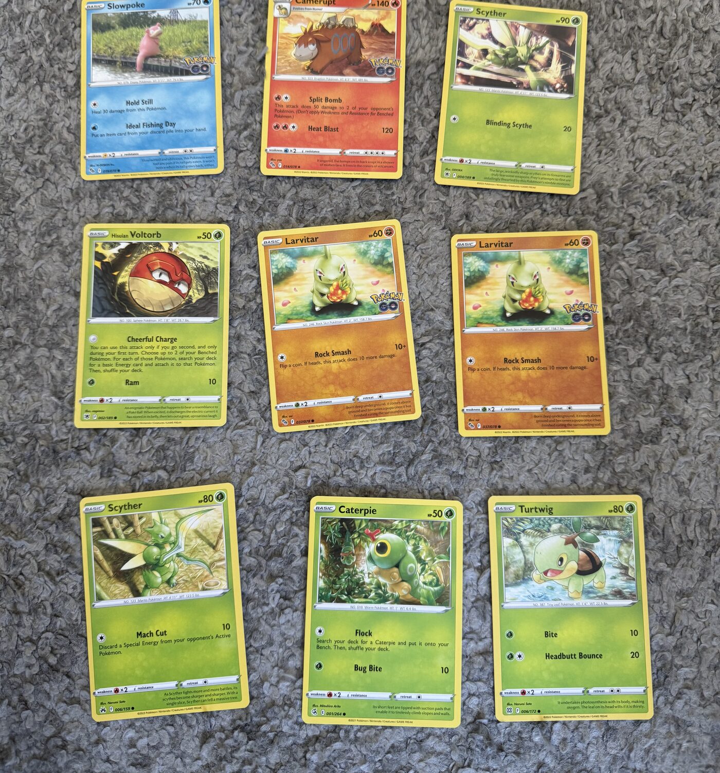 Nine Pokémon cards laid out in a grid on a grey rug, ready to be scanned in one photo.